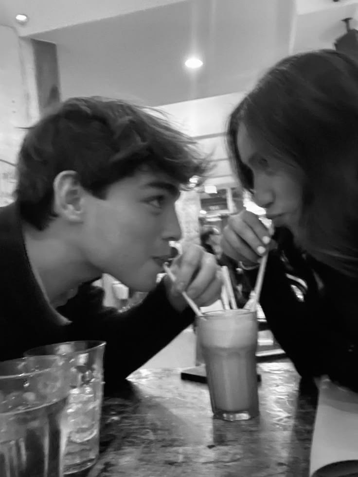 A black-and-white photograph of a young couple sharing a milkshake across a café counter.