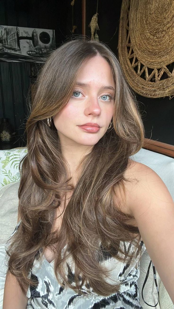 A young woman with long brunette hair and blue eyes looks toward the camera in warm indoor light.